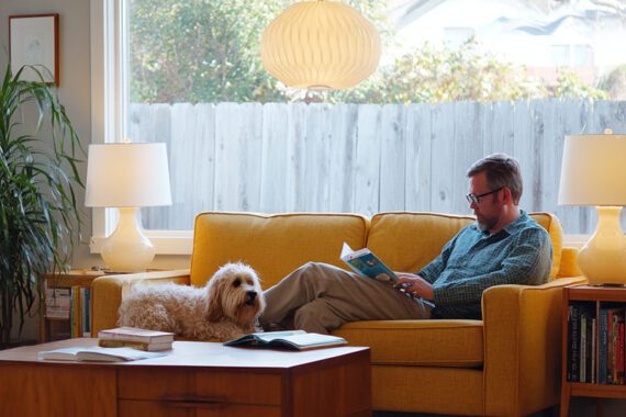 A man reading a book on a couch next to a dog A man reading a book on a couch next to a dog