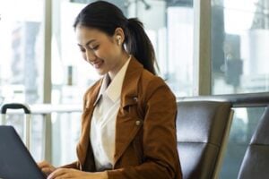 Smiling woman sitting in an airport lounge, wearing wireless earbuds and a brown jacket, working on a laptop with luggage beside her.