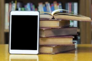 Photo of an ebook reader next to a stack of print books on a table