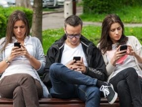 Photo of 2 females and 1 male on a park bench looking at their phones