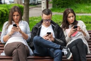 Photo of 2 females and 1 male on a park bench looking at their phones